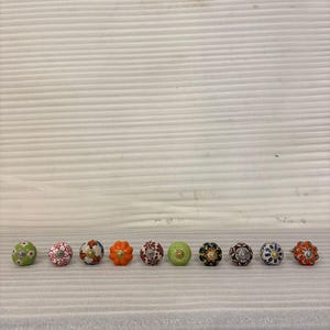 Könnte beinhalten: Eine Sammlung von zehn dekorativen Keramik-Schrankknöpfen. Die Knöpfe weisen verschiedene Farben und Muster auf, darunter florale Designs, Punkte und geometrische Formen. Jeder Knopf ist ein einzigartiges Akzentstück für Möbel.