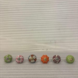 以下が含まれることがあります： 6つの装飾的なセラミックキャビネットノブのコレクション。ノブは丸く、花柄やオレンジ色の花の形など、さまざまな色と模様が特徴です。各ノブには金属製のベースが付いています。