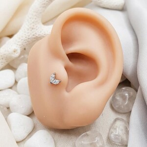 May include: A silver-toned ear piercing stud featuring two marquise-cut clear stones arranged in a leaf-like shape. The jewellery is positioned in the tragus area of a neutral-toned ear model, showcasing the metallic setting and faceted stones.