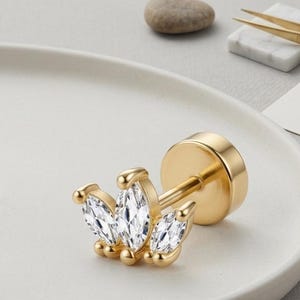 Pode incluir: Um brinco de pino com parte traseira plana em tom dourado, apresentando um design de coroa com três pedras transparentes de corte marquise. O brinco possui uma haste reta e um fecho circular plano com um acabamento metálico polido.