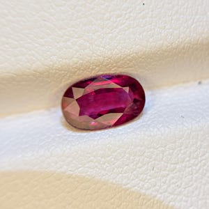 May include: A faceted oval-shaped gemstone with a deep, vibrant red color. The stone features a polished surface with multiple geometric facets that reflect light, creating a brilliant appearance. The gemstone is displayed against a textured white surface.