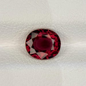 May include: A deep red, oval-cut gemstone with a faceted surface. The stone displays a rich, saturated crimson hue with sharp, geometric light reflections across its polished top, showcasing a clear, symmetrical arrangement of facets.