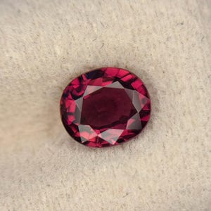 Puede incluir: Una piedra preciosa de corte ovalado con un color rojo profundo y saturado y una superficie multifacetada. La piedra muestra una calidad brillante y reflectante con facetas geométricas nítidas que captan la luz, mostrando un tono carmesí.