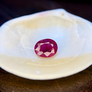 以下が含まれることがあります： 深い赤色で内部の透明度が見える、ファセットカットされた楕円形のルビーの宝石。滑らかで白い凹型の貝殻の中に置かれており、宝石の鮮やかな赤く磨かれた表面と鮮明なコントラストを生み出しています。