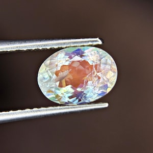 Könnte beinhalten: Ein oval geschliffener Edelstein mit facettierter Oberfläche, der irisierende Lichtreflexe in Orange-, Blau- und Weißtönen zeigt. Der Stein wird von einer silberfarbenen Metallpinzette gehalten und zeigt seine durchscheinende Beschaffenheit.