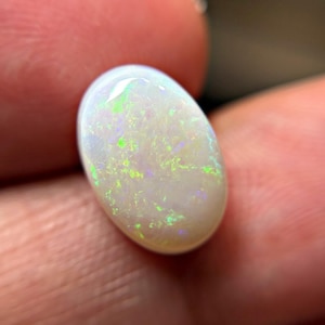 May include: An oval-shaped white opal gemstone featuring a smooth, polished surface with internal flashes of vibrant neon green and subtle violet light play throughout the stone, showcasing a translucent quality and a high-gloss finish.