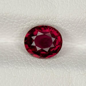 May include: A deep red, oval-cut gemstone featuring a faceted surface with a central star-like pattern. The stone displays a rich, saturated crimson hue with internal reflections and sharp geometric edges throughout its polished structure.