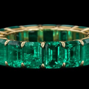 May include: A gold ring featuring a continuous band of emerald-cut emeralds. The vibrant green gemstones are set in a gold band, creating a luxurious and elegant design. The ring is displayed against a black background.