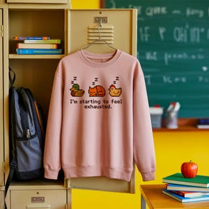 Peut inclure: Un sweat-shirt rose clair à col rond. Il présente un motif pixélisé d'un canard, d'un chat et d'une poule endormis, avec le texte "I'm starting to feel exhausted."