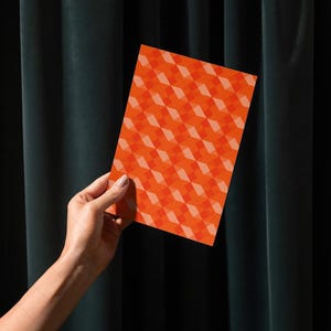Puede incluir: Una tarjeta rectangular con un patrón geométrico repetitivo de formas de diamante naranja y naranja claro. La tarjeta se sostiene contra un fondo de cortina verde oscuro.