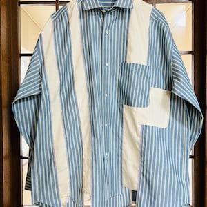 Pode incluir: Camisa abotoada com riscas verticais em azul claro e branco. A camisa apresenta um painel creme na frente e um bolso a condizer. A camisa está pendurada num cabide de madeira.