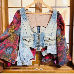May include: A distinctive denim vest with bell sleeves. The vest has a light blue wash, a button-up front, and two pockets. The sleeves are made of a colourful patterned fabric with red, blue, and yellow hues.