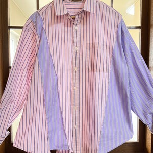 Puede incluir: Una camisa abotonada con diseño de patchwork. Presenta rayas verticales rosas y lavanda, un bolsillo en el pecho y mangas largas. La camisa está colgada en una percha de madera.