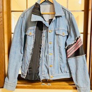 May include: Light blue denim jacket with gold-colored buttons and a black satin panel down the front. The right sleeve features a black patch with pink stripes. The jacket is hanging on a wooden hanger.