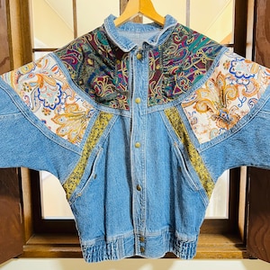Puede incluir: Chaqueta vaquera vintage con un diseño patchwork único. La chaqueta presenta mezclilla azul claro con acentos de tela estampada de cachemira y floral en tonos naranja, azul y verde. Tiene cuello, cierre de botones y bolsillos.