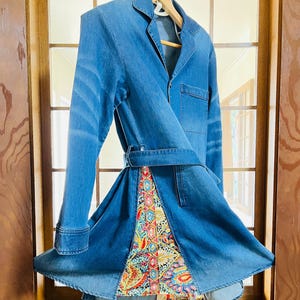 May include: A blue denim jacket with a unique design. The jacket features a belt and a colourful patterned fabric insert at the bottom. The jacket is hanging on a wooden hanger in front of a window.