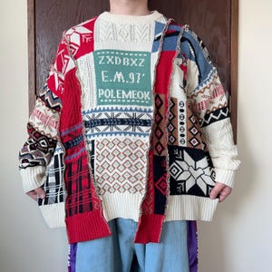 Op de afbeelding: Een patchwork trui in crème, rood, zwart en blauw. De trui heeft verschillende geometrische patronen, waaronder sneeuwvlokken en ruiten, en de tekst "ZXDBXZ E.M.97' POLEMEOK". De trui heeft een ronde hals en lange mouwen.