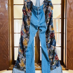 Może przedstawiać: Jasnoniebieskie dżinsy z bocznymi panelami z tkaniny we wzory kwiatowe. Kwiatowy wzór przedstawia czerwone, żółte i białe kwiaty na niebieskim tle. Dżinsy wiszą przed oknem.