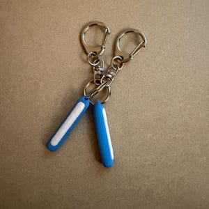 May include: Two keychains with silver metal clasps and blue, cylindrical charms. Each charm has a white stripe down the center. The keychains are displayed against a neutral, tan background.
