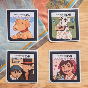 May include: Four Nintendo DS game cartridge stickers. The stickers feature colorful illustrations from popular games: Nintendogs, Animal Crossing, Professor Layton, and Cooking Mama. Each sticker has a black border and the Nintendo DS logo.