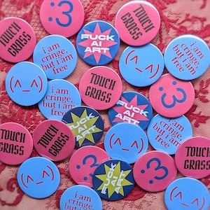 May include: A collection of circular pin-back buttons in pink and blue featuring various designs including the text "TOUCH GRASS", "FUCK AI ART", "i am cringe, but i am free", and emoticons like ":3" and "(^_^")" printed in contrasting colours.