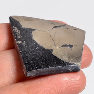 Puede incluir: Un espécimen mineral trapezoidal pulido que presenta un contraste marcado entre una sección de pirita metálica de color latón y una sección de piedra negra mate. La superficie muestra fisuras naturales y un acabado plano y liso.