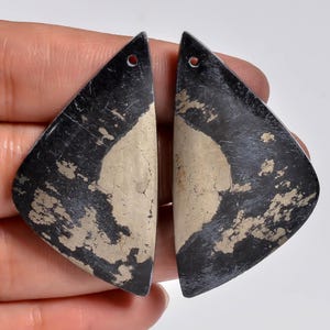 Puede incluir: Dos colgantes de forma triangular hechos de piedra negra con inclusiones naturales de pirita de color oro metálico. Cada pieza presenta una superficie lisa y pulida y un pequeño orificio circular perforado cerca de la punta superior.