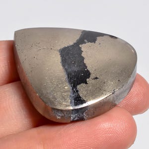 May include: A polished, triangular-shaped piece of pyrite with a metallic, brassy gold lustre. The stone features a distinct, irregular dark black vein running vertically through the centre, contrasting with the reflective, textured surface.
