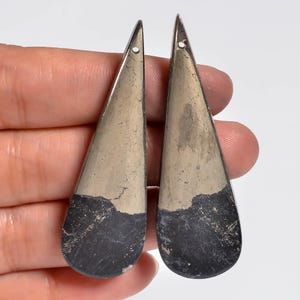 May include: Two elongated, teardrop-shaped pendants featuring a dual-tone design. The upper section is a metallic, reflective gold-toned pyrite, while the lower portion consists of a matte, dark charcoal-grey stone with visible natural texture.