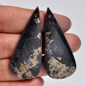 May include: Two teardrop-shaped pendants made of dark grey stone with visible metallic gold-coloured inclusions. Each pendant features a smooth, polished surface and a small circular hole drilled near the pointed top for suspension purposes.