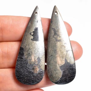 Puede incluir: Dos colgantes en forma de lágrima hechos de pirita pulida con inclusiones naturales de color gris oscuro. Cada pieza presenta una superficie metálica de color oro latón con manchas negras irregulares y un pequeño orificio en la punta.