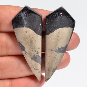 Puede incluir: Dos colgantes en forma de escudo hechos de piedra negra y dorada metálica. Cada pieza presenta una parte superior puntiaguda con un pequeño orificio perforado para colgar, mostrando una superficie lisa y pulida con variaciones de color.