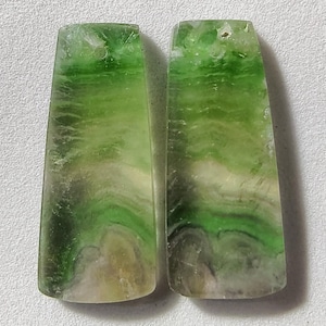 May include: Two polished, rectangular gemstones with a trapezoid shape. The stones feature a mix of green, white, and tan colors, with a swirling, layered pattern. The gemstones are set against a neutral background.