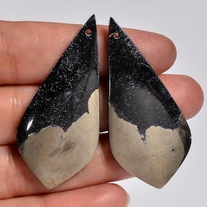 May include: Two teardrop-shaped pendants featuring a polished black surface on the upper portion and a contrasting matte beige, textured mineral section on the lower half. Each piece includes a small circular hole at the top for suspension.