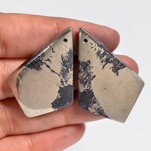 Puede incluir: Dos colgantes de pirita asimétricos en forma de cometa con una superficie metálica dorada reflectante e inclusiones gris oscuro mate. Cada pieza presenta un pequeño orificio circular en la esquina superior para colgar, mostrando una textura mineral.