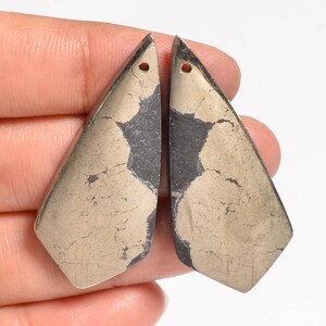 Puede incluir: Dos colgantes de pirita en forma de lágrima con una superficie moteada de color dorado metálico y gris oscuro. Cada pieza presenta un acabado liso y pulido, con un pequeño orificio perforado en la punta superior para colgar.