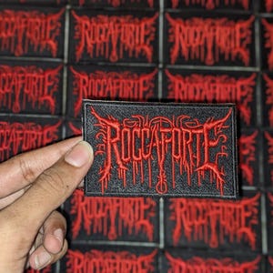 Op de afbeelding: Zwart rechthoekig embleem met het woord "ROCCAFORTE" geborduurd in rode, gotische letters. De patch wordt voor een achtergrond van vergelijkbare patches gehouden. Het ontwerp heeft een heavy metal esthetiek.