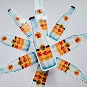 May include: A collection of Topo Chico water bottle stickers. The stickers feature a watercolor design of the bottle with a blue and white color scheme. The label reads "Topo Chico Agua Mineral" in orange and black.