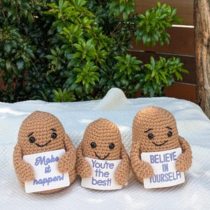 May include: Three brown crocheted potato-shaped figures with smiling faces, each holding a white sign with motivational phrases in purple text: "Make it happen!", "You're the best!", and "Believe in yourself". The figures are on a white surface.