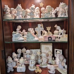 May include: A collection of numerous matte porcelain figurines displayed on three glass shelves within a wooden cabinet. The figurines feature children with teardrop-shaped eyes, animals, and decorative items in soft pastel colors and white.