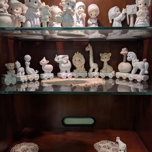 May include: A collection of matte porcelain figurines displayed on two shelves. The top shelf features various children in winter clothing. The middle shelf holds a train of animal figurines including an elephant, lion, giraffe, and horse.