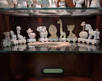 May include: A collection of matte porcelain figurines displayed on two shelves. The top shelf features various children in winter clothing. The middle shelf holds a train of animal figurines including an elephant, lion, giraffe, and horse.