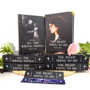 Puede incluir: Una colección de libros con cubiertas negras. Los títulos son "All The Wrong Things" y "The Right Thing To Do". Las cubiertas presentan ilustraciones de personas y el texto "Book One" y "Book Two".