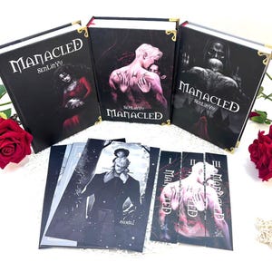 Op de afbeelding: Drie hardcover boeken en verschillende kaarten met illustraties. De boeken hebben zwarte covers met gouden hoekaccenten en de titel "Manacled" in het wit. De kaarten bevatten zwart-wit illustraties. De boeken en kaarten zijn gerangschikt op een wit oppervlak.