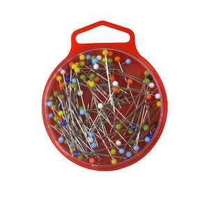 Puede incluir: Un recipiente redondo de plástico rojo con una pestaña para colgar, lleno de una colección de alfileres de costura con pequeñas cabezas esféricas multicolores en tonos azul, amarillo, verde, naranja y blanco, dispuestos al azar.