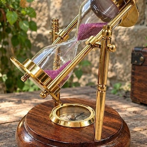 May include: A rotating hourglass with a polished brass frame and clear glass bulbs containing vibrant purple sand. The hourglass is mounted on a circular dark brown wooden base that features an integrated brass compass in the center.