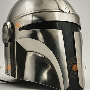 Op de afbeelding: Een zilveren Mandalorian helm met een afgeronde bovenkant en een T-vormig vizier. De helm heeft een donkergrijze frontplaat en is waarschijnlijk gemaakt van metaal. Het ontwerp doet denken aan een ruimtethema-personage.