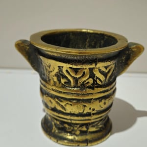 Peut inclure: Un petit mortier et pilon en laiton antique avec une patine sombre. Le mortier a une large ouverture, deux poignées et est décoré de motifs en relief ornés. La base est également décorée de motifs en relief.
