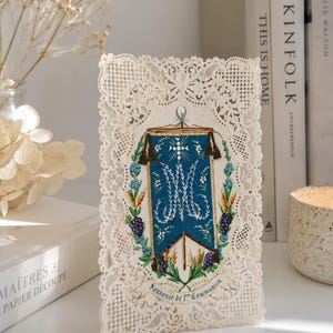 Peut inclure: Une carte rectangulaire avec une bordure en dentelle crème complexe entourant une illustration centrale d'une bannière bleue. La bannière affiche un monogramme blanc, des motifs floraux et le texte Souvenir de 1re Communion en bas.