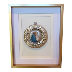 Peut inclure: Une plaque murale circulaire encadrée présentant un portrait central peint d'une personne en châle bleu tenant un enfant. La plaque possède une bordure métallique ornée de détails floraux en relief dans un cadre rectangulaire doré.